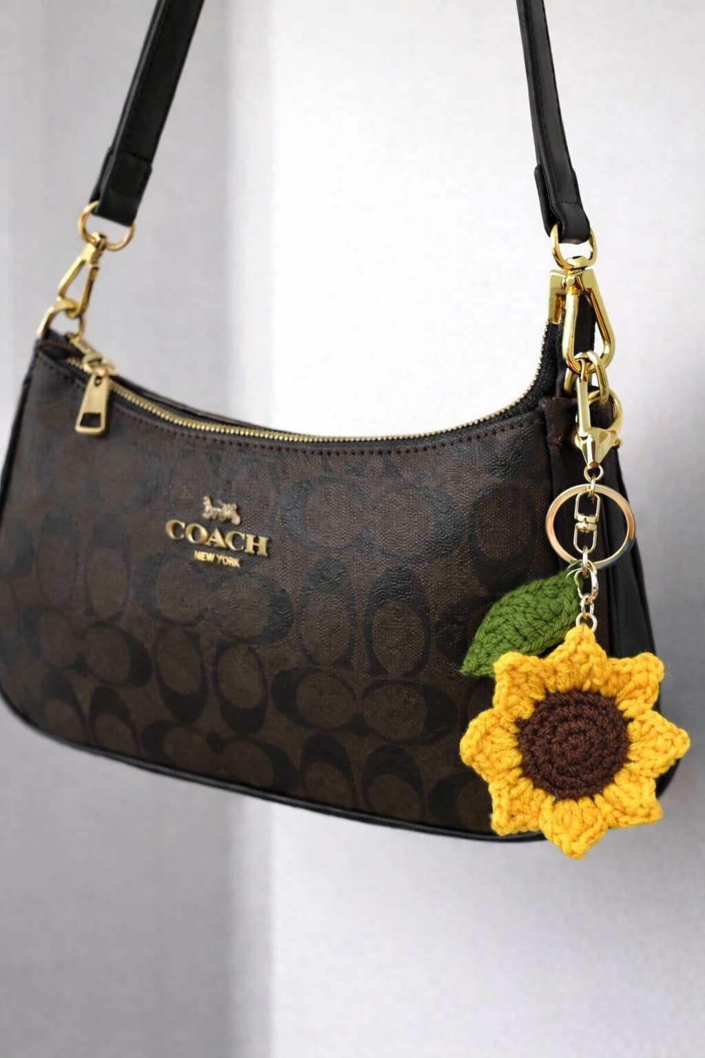 Hand holding a large crochet sunflower keychain with a green leaf charm and silver clasp on coach bag.